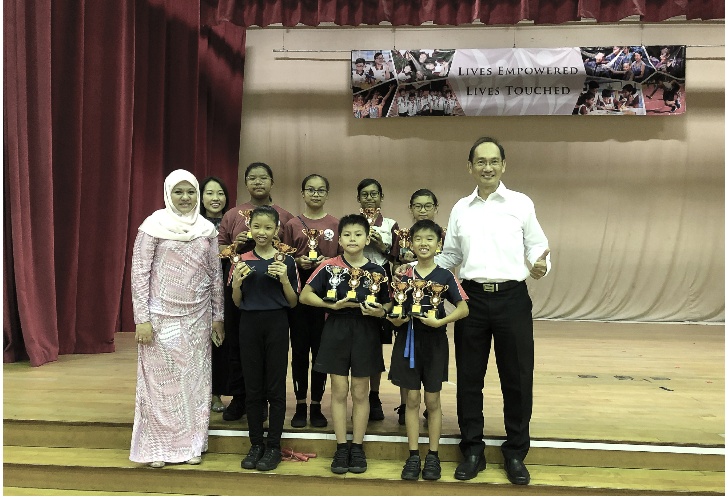 Group of children holding trophies posing with two adults on a school stage under a "Lives Empowered, Lives Touched" banner.